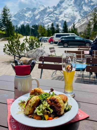 Ein Teller mit gebratenen Kräutern und ein Bier, im Hintergrund Berge und Bäume.