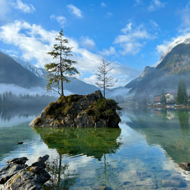 Ruhiger See, Hintersee, mit Felseninsel und zwei Bäumen, umgeben von Bergen und Nebel.