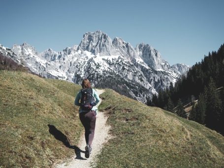 Eine Frau mit Rucksack wandert auf einem Pfad auf der Bindalm, in bergiger Landschaft mit schneebedeckten Gipfeln.