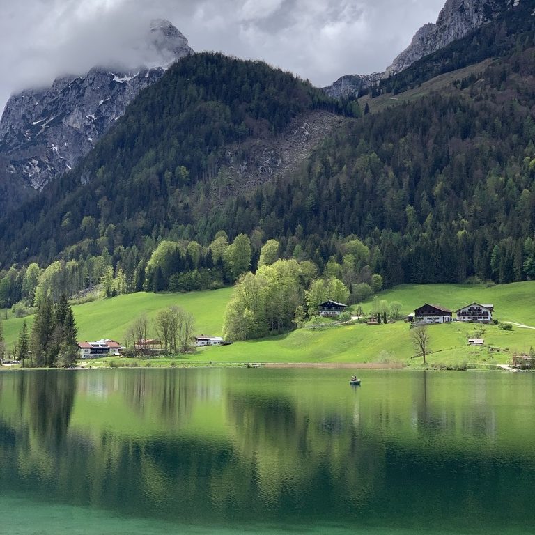Landschaft mit Bergen, grünen Wiesen und einem ruhigen See, Hintersee in Ramsau bei Berchtesgaden, unter bewölktem Himmel.