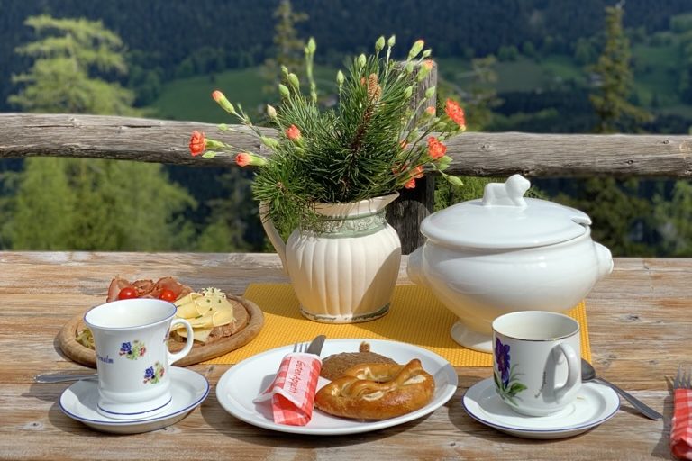 Einkehr mit Tee, Kaffee, Weisswurst und Brezen und einer blühenden Pflanze auf einem Holztisch auf der Schärtenalm.