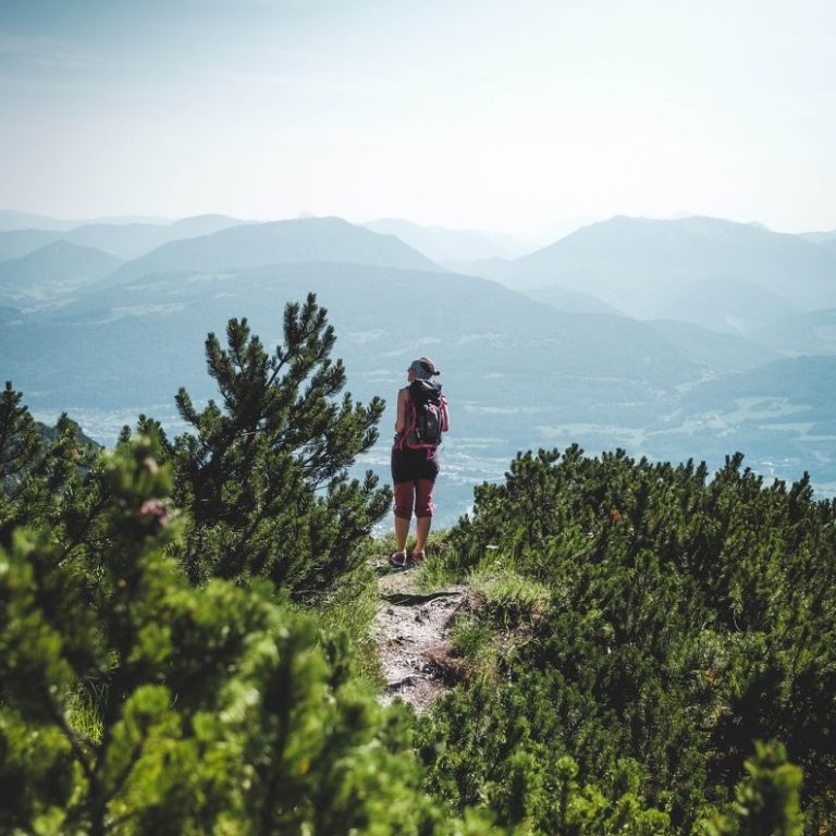 Frau mit Rucksack steht auf einem Bergweg am Untersberg, umgeben von Pflanzen und Bergen im Hintergrund.