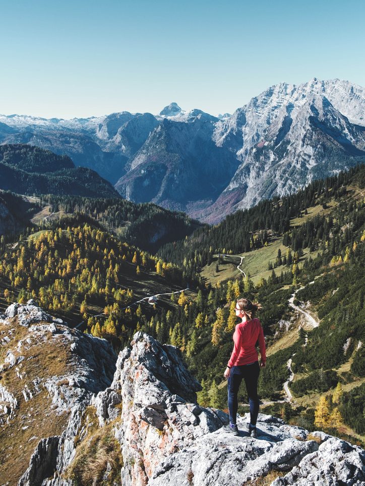Eine Frau steht auf einem Gipfel im Nationalpark Berchtesgaden und blickt auf eine bergige Landschaft im Herbst. 