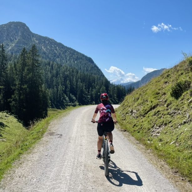 Fahrradfahrerin auf einem Schotterweg auf dem Weg zur Kallbrunnalm, umgeben von Bergen und Bäumen.
