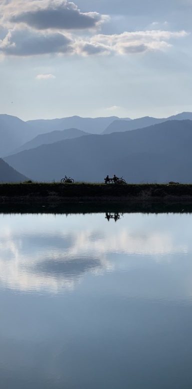 Zwei Radfahrer sitzen am Ufer eines ruhigen Speicherteichs im Nationalpark Berchtesgaden, umgeben von Bergen und Wolken.