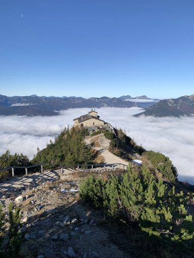 Berggipfel mit einer Hütte, umgeben von Nebel und Bergen im Hintergrund.