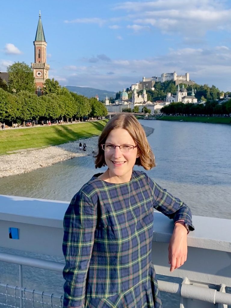 Natalia Herzinger in Salzburg Junge Frau in einem karierten Kleid steht am Fluss mit einer Stadt im Hintergrund.